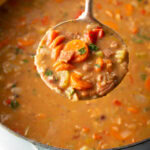 Top down view soup made with 15 beans and veggies in a large cooking pot. A metal ladle is showing a spoonful to camera.