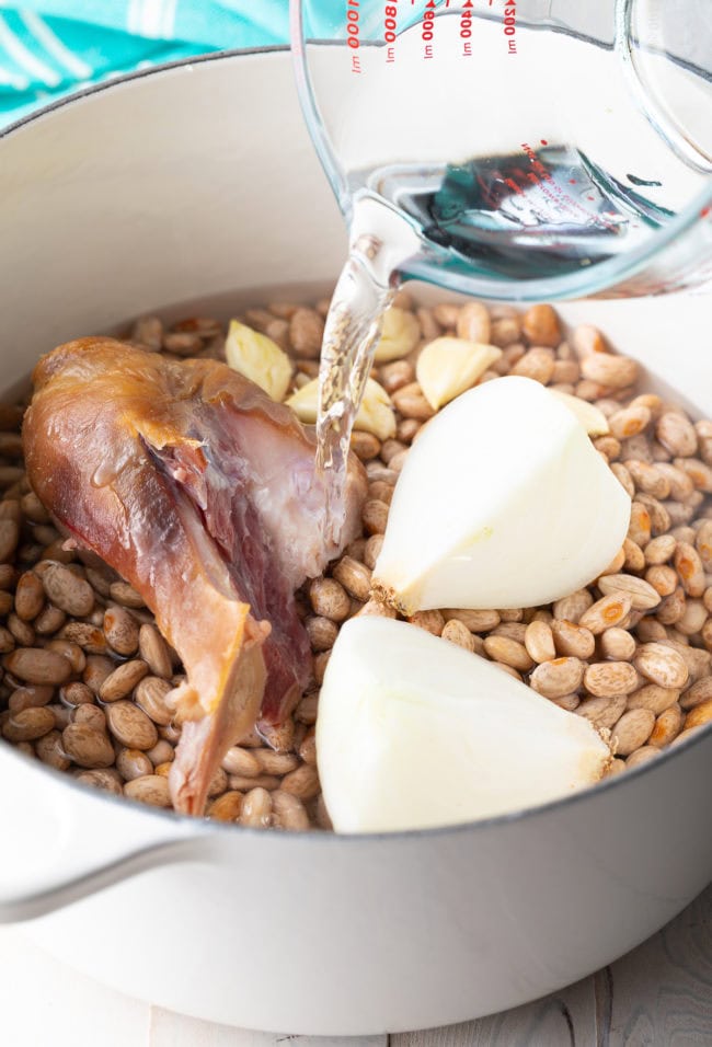 Pouring water into a dutch oven full of beans, onion, garlic and ham hock. 