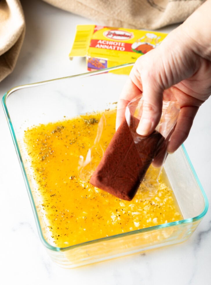 Hand adding a block of achiote paste to a yellow mixture in a square glass dish.