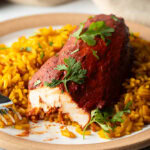 Plate of yellow rice and achiote chicken breast. A fork has take a bite from the chicken to show the inner meat