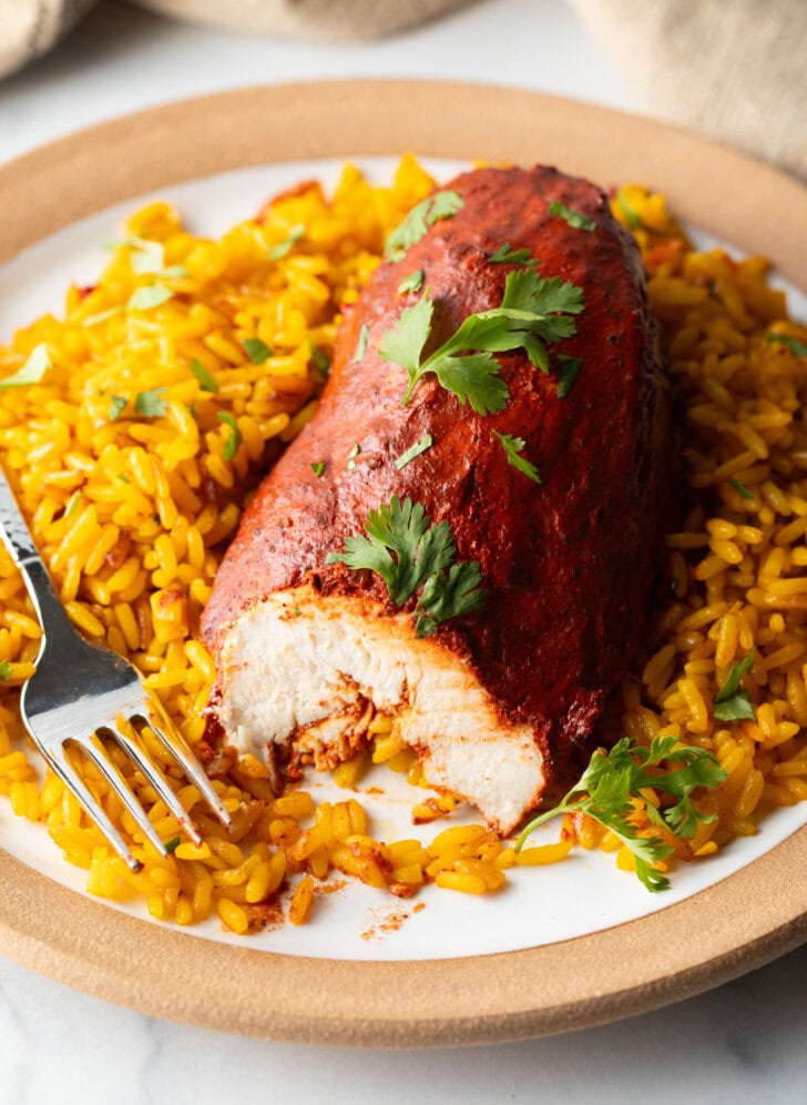 Plate of yellow rice and achiote chicken breast. A fork has take a bite from the chicken to show the inner meat