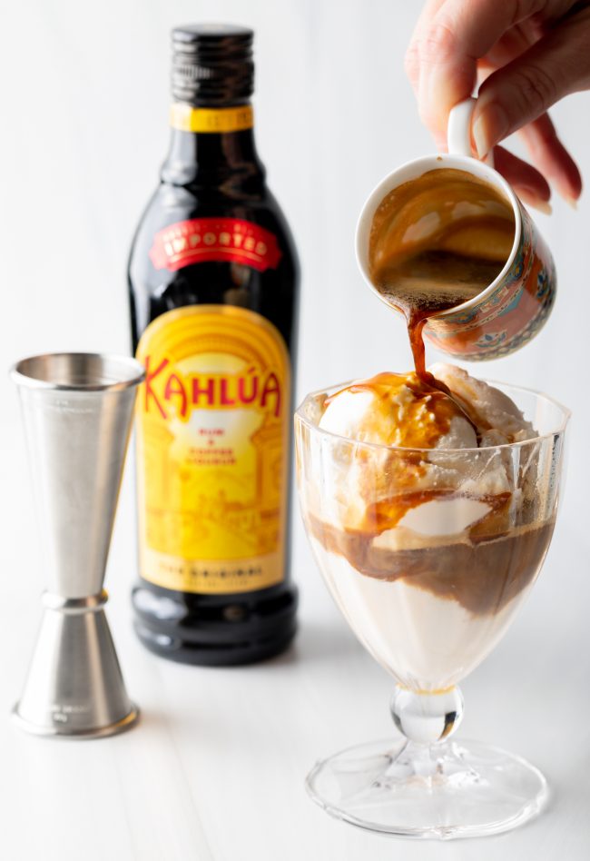 Hand pouring espresso from a small mug over a glass of ice cream and Kahlua.