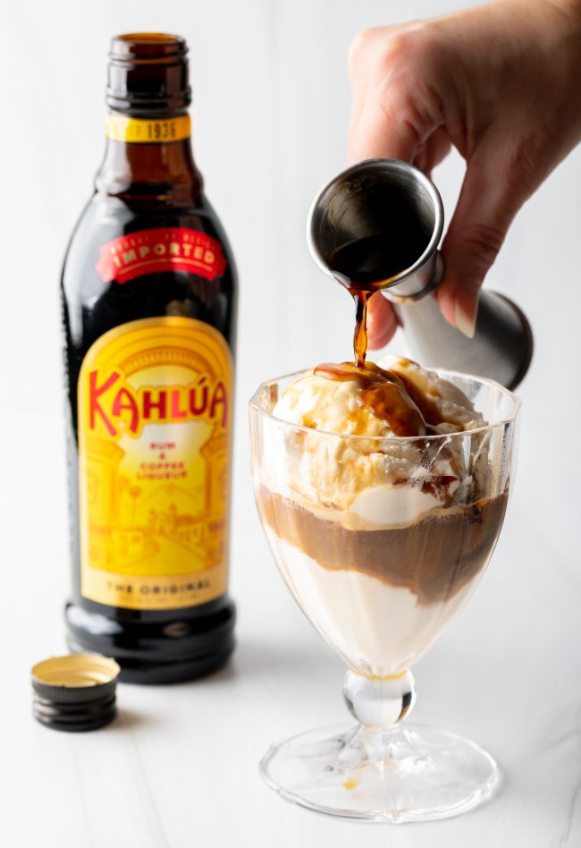 Hand pouring Kahlua from a metal jig into a glass with ice cream.