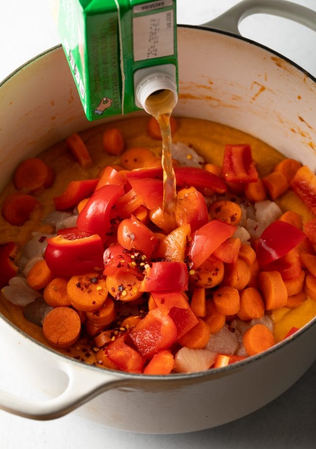 Pouring chicken broth over chopped peppers and carrots with chicken and peanut puree in a large cooking pot.