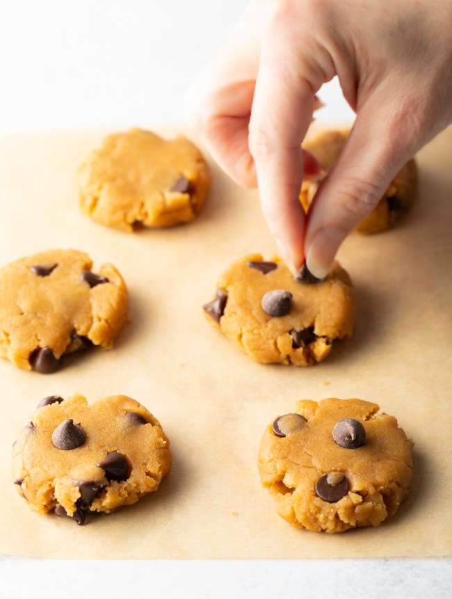 Hand adding chocolate chips to flattened raw cookie dough on parchment.