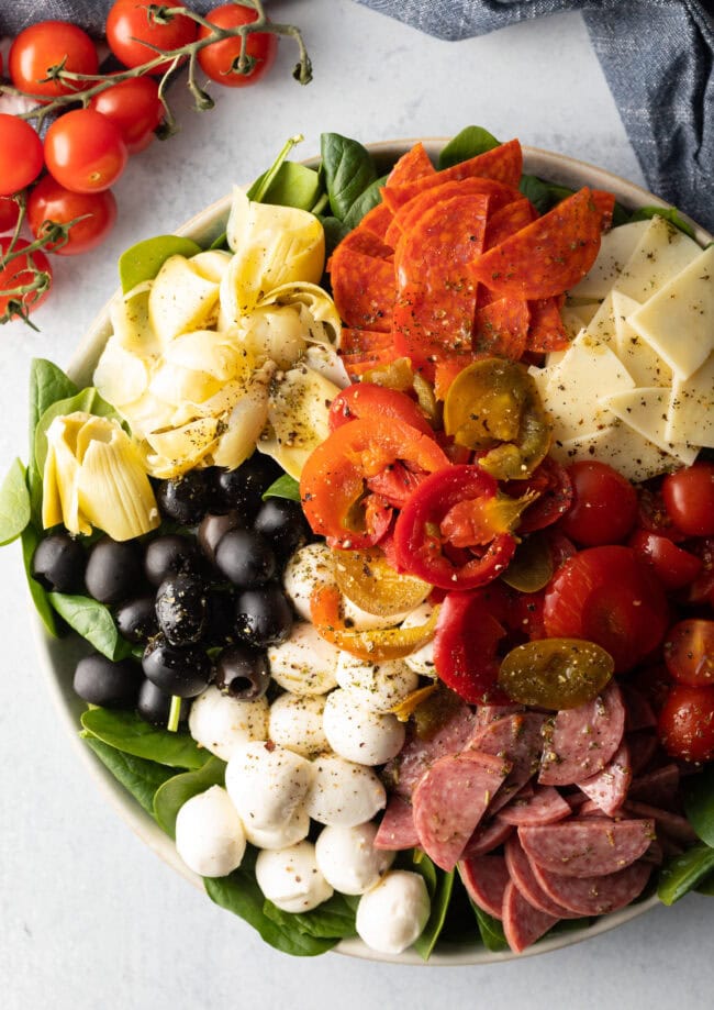 Top down view of antipasto salad, loaded with spinach, black olives, mozzarella balls, pepperoni slices, sausage slices, cherry tomatoes, cheese slices, artichoke hearts, and pickled peppers.