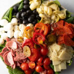 Top down view of antipasto salad, loaded with spinach, black olives, mozzarella balls, pepperoni slices, sausage slices, cherry tomatoes, cheese slices, artichoke hearts, and pickled peppers.