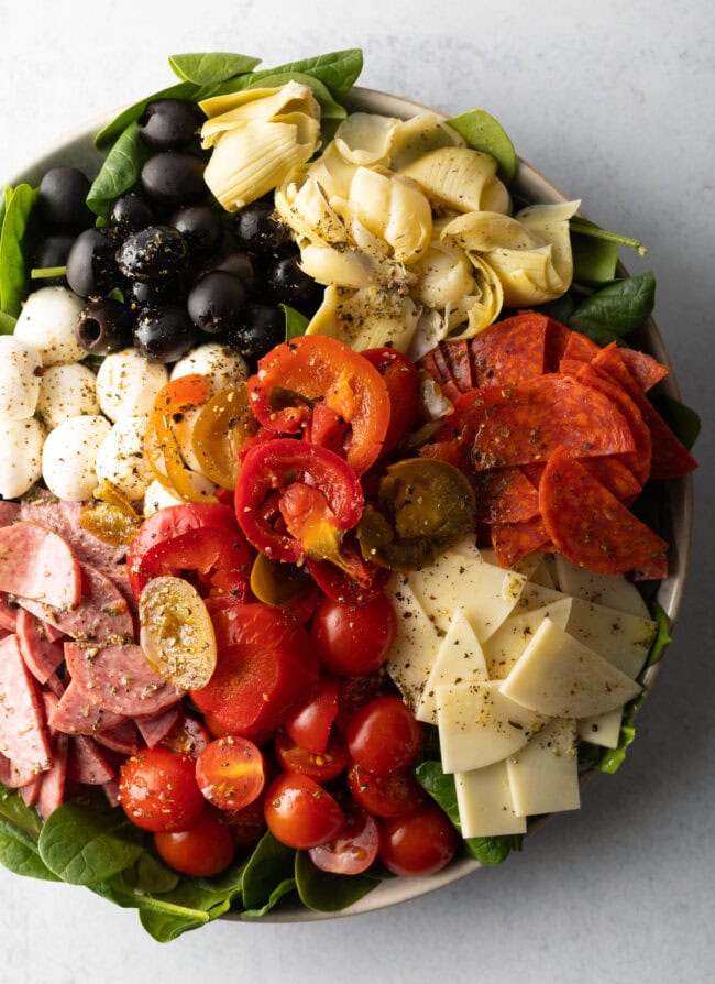 Top down view of antipasto salad, loaded with spinach, black olives, mozzarella balls, pepperoni slices, sausage slices, cherry tomatoes, cheese slices, artichoke hearts, and pickled peppers.