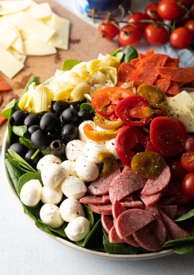 Top down view of antipasto salad, loaded with spinach, black olives, mozzarella balls, pepperoni slices, sausage slices, cherry tomatoes, cheese slices, artichoke hearts, and pickled peppers.