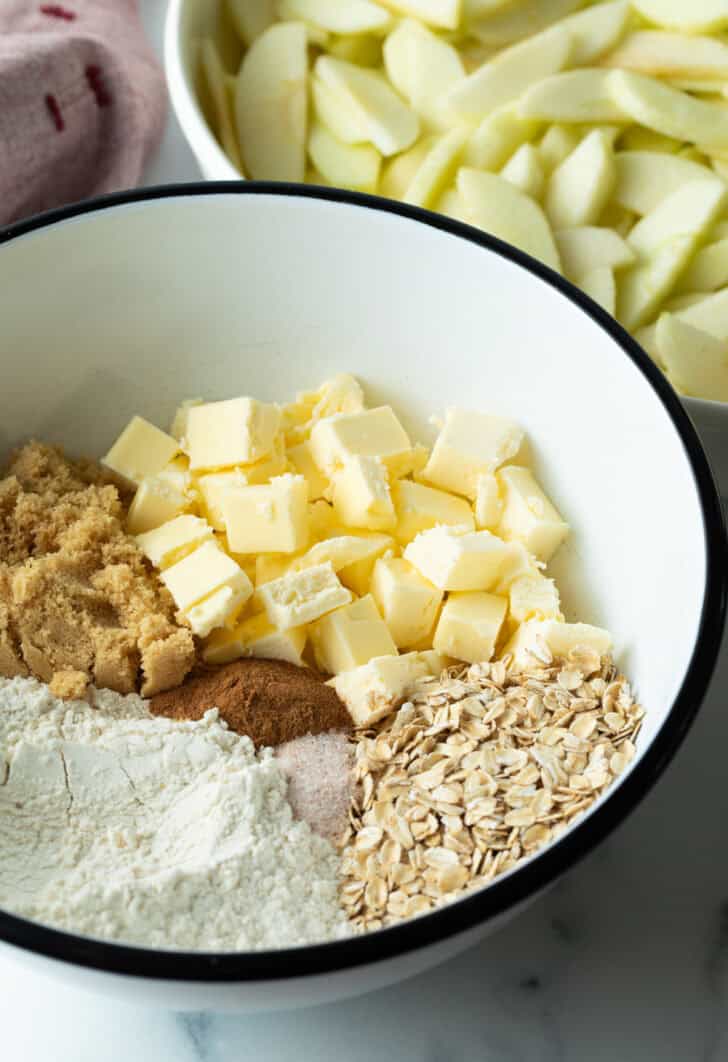 White bowl with flour, cubes of butter, brown sugar, oats, and spices.