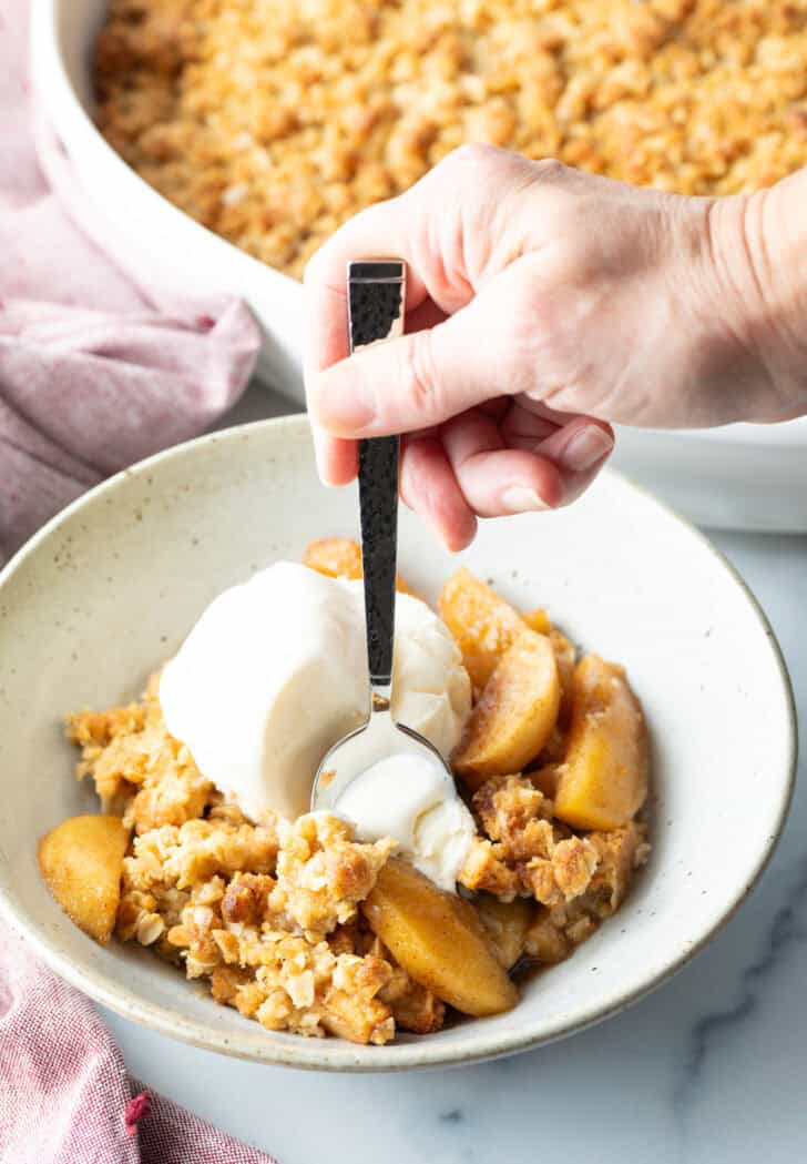 White bowl with golden apple crisp topped with a big scoop of ice cream. A hand with a metal spoon taking a bite of ice cream.