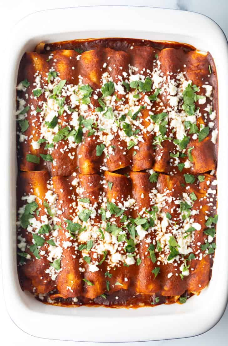 Large white rectangle baking dish with a dozen rolled enchiladas covered in red sauce, sprinkled with cotija cheese and chopped cilantro.