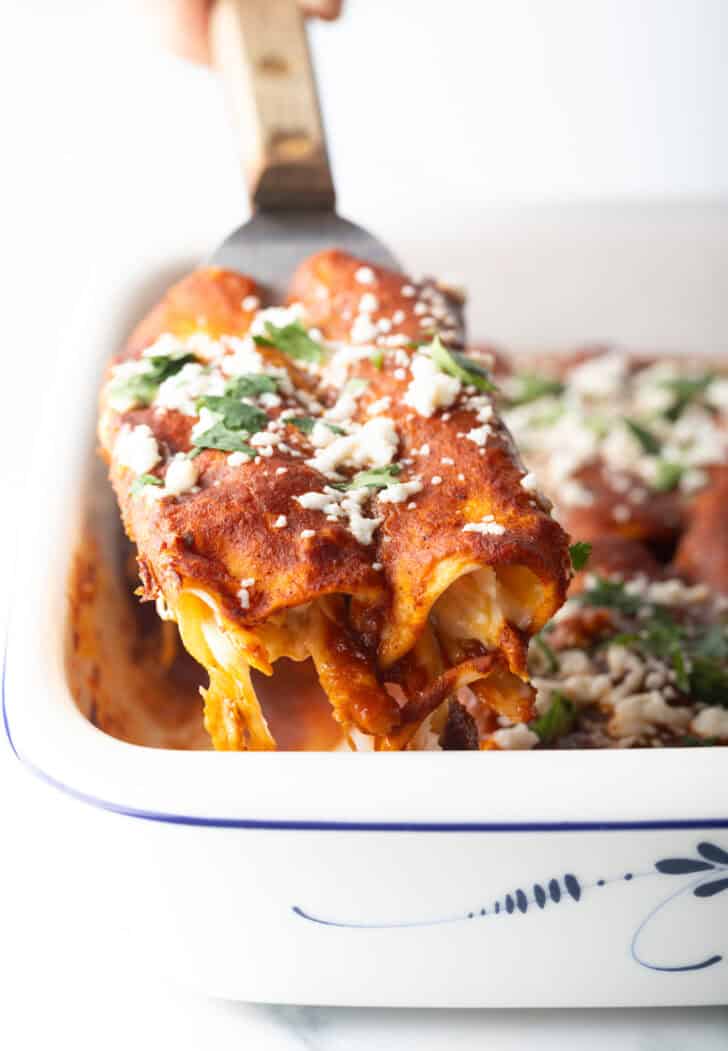 Metal spoon lifting two enchiladas from the baking dish.