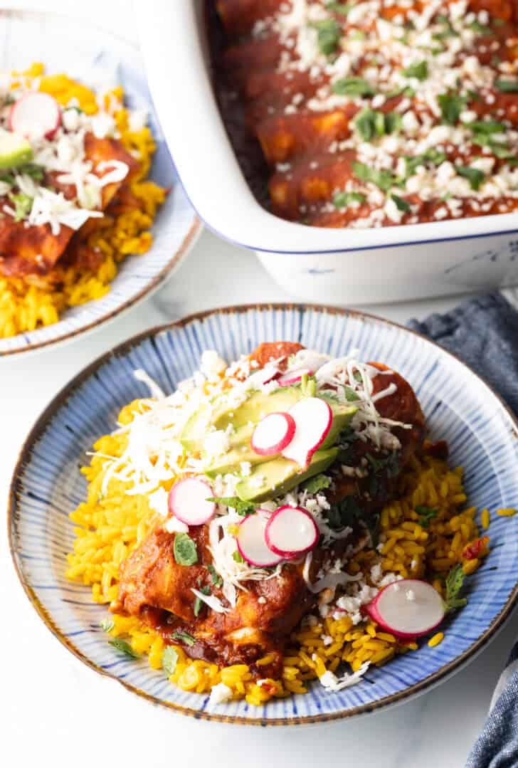 Large plate with 2 enchiladas covered in red sauce, on a pile of fluffy yellow rice and topped with fresh cabbage slaw, sliced radishes, and avocado.