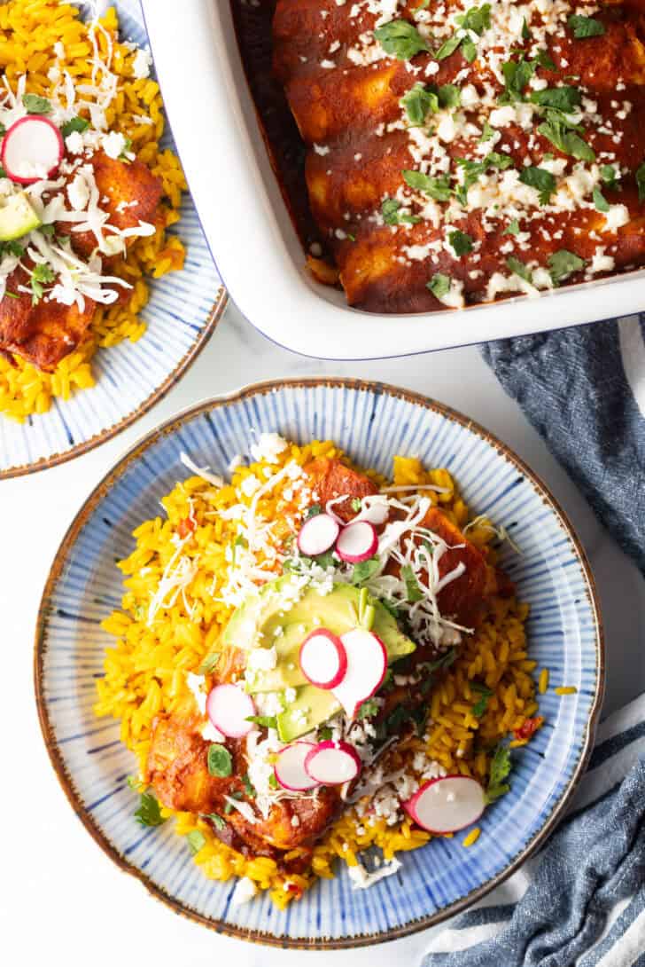 Large plate with 2 authentic enchiladas covered in red sauce, on a pile of fluffy yellow rice and topped with fresh cabbage slaw, sliced radishes, and avocado.