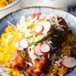 Large plate with 2 enchiladas covered in red sauce, on a pile of fluffy yellow rice and topped with fresh cabbage slaw, sliced radishes, and avocado.