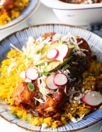 Large plate with 2 enchiladas covered in red sauce, on a pile of fluffy yellow rice and topped with fresh cabbage slaw, sliced radishes, and avocado.