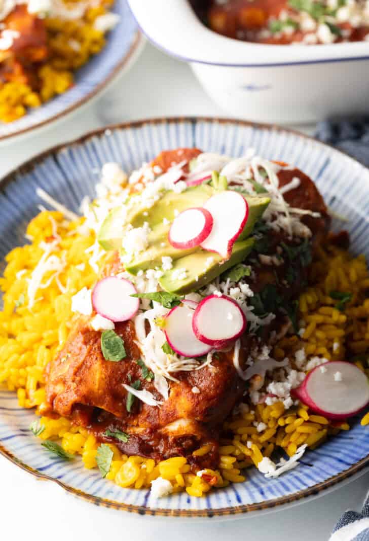 Authentic Enchilada Recipe - Large plate with 2 enchiladas covered in red sauce, on a pile of fluffy yellow rice and topped with fresh cabbage slaw, sliced radishes, and avocado.