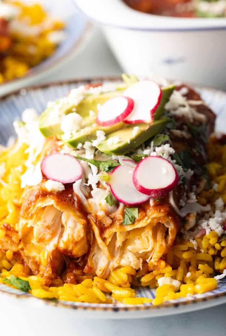 Large plate with 2 enchiladas covered in red sauce, on a pile of fluffy yellow rice and topped with fresh cabbage slaw, sliced radishes, and avocado.