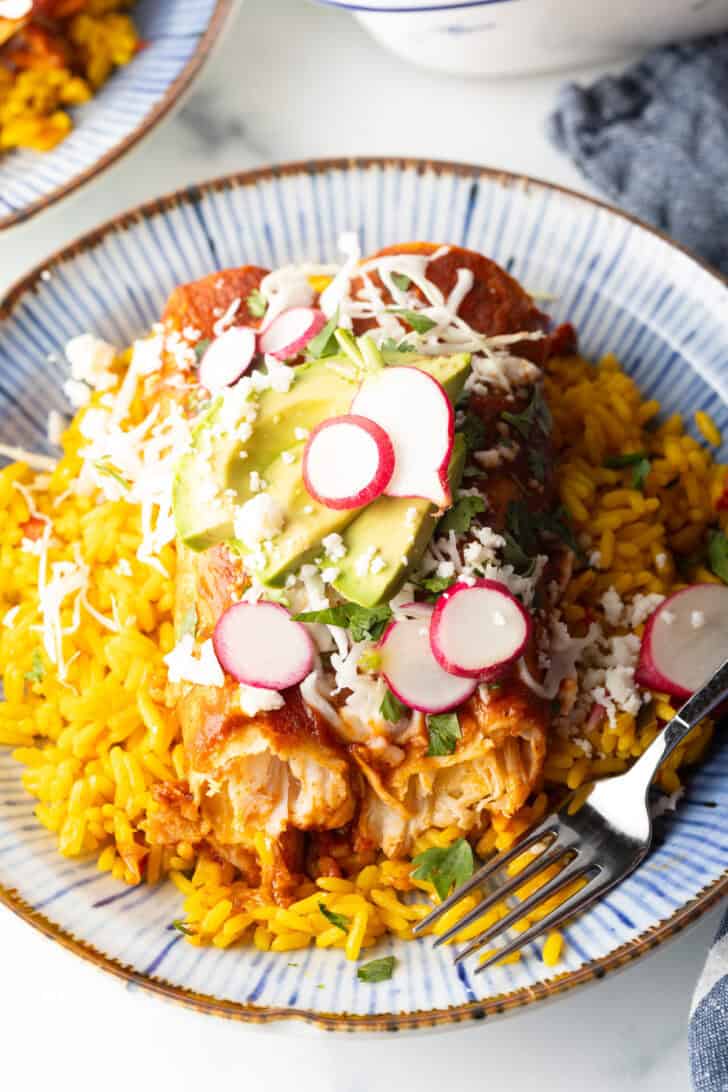 Large plate with 2 enchiladas covered in red sauce, on a pile of fluffy yellow rice and topped with fresh cabbage slaw, sliced radishes, and avocado.