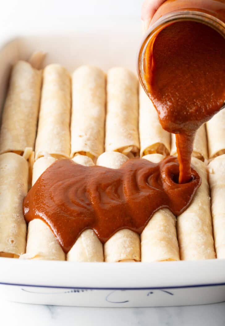 Pouring red sauce from a glass jar over the top of a dozen rolled enchiladas in a white baking dish.