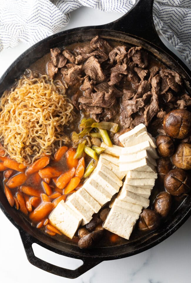 Deep black skillet with cooked beef sukiyaki loaded with tofu, steak, carrots, mushroom, green beans, and bean sprouts with authentic sauce.