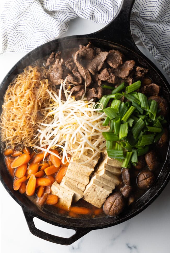 Deep black skillet with cooked beef sukiyaki loaded with tofu, steak, carrots, mushroom, green beans, and bean sprouts with authentic sauce.