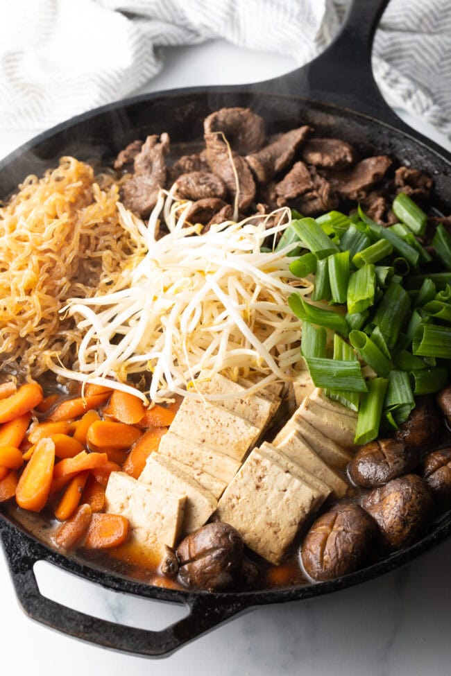 Deep black skillet with cooked beef sukiyaki loaded with tofu, steak, carrots, mushroom, green beans, and bean sprouts with authentic sauce.