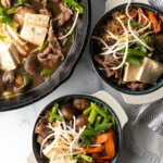 Top down 2 bowls of beef sukiyaki loaded with tofu, steak, carrots, mushroom, green beans, and bean sprouts.