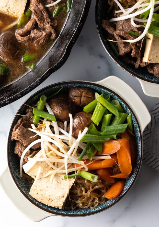 Top down 2 bowls of beef sukiyaki loaded with tofu, steak, carrots, mushroom, green beans, and bean sprouts.