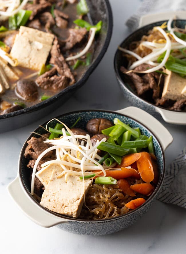 Bowls of beef sukiyaki loaded with tofu, steak, carrots, mushroom, green beans, and bean sprouts.