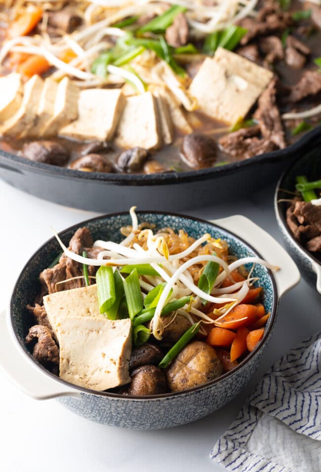 Bowls of beef sukiyaki loaded with tofu, steak, carrots, mushroom, green beans, and bean sprouts.