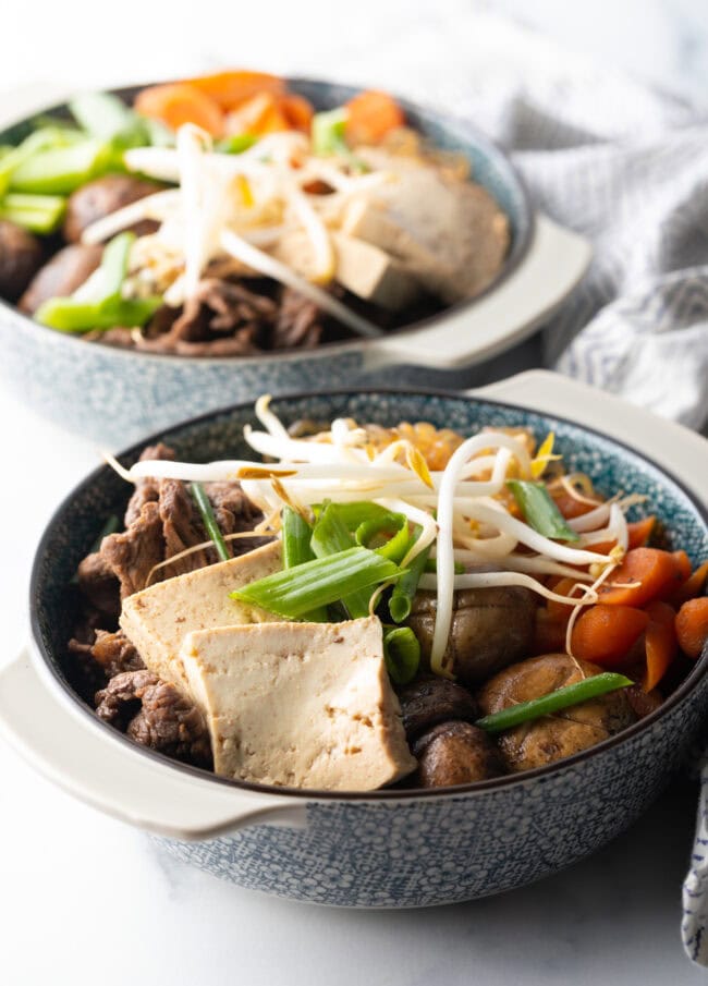 Bowls of beef sukiyaki loaded with tofu, steak, carrots, mushroom, green beans, and bean sprouts.