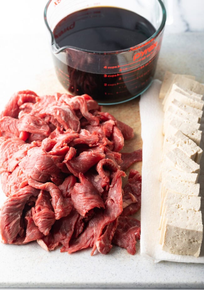 Thin strips of raw beef, block of tofu, and glass measuring cup with sauce.