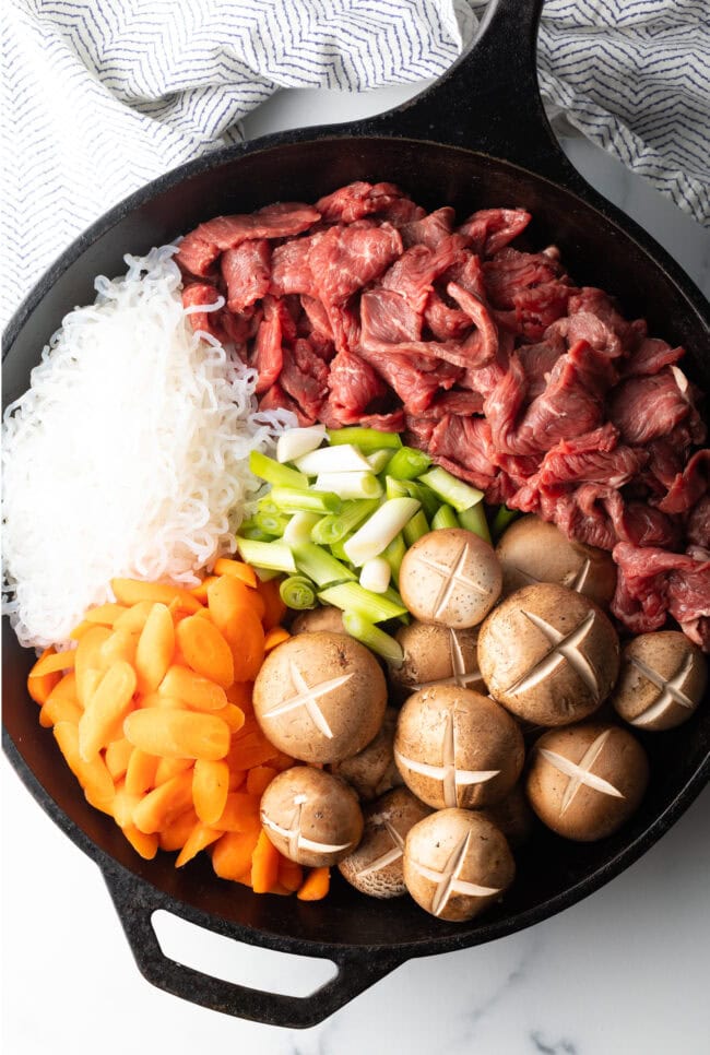 Raw recipe ingredients in a large black skillet: Raw beef, noodles, mushrooms, carrots, and beans.