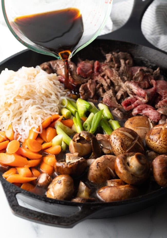 Pouring sukiyaki sauce into a skillet with sections of noodles, mushrooms, beef, carrots, and beans.