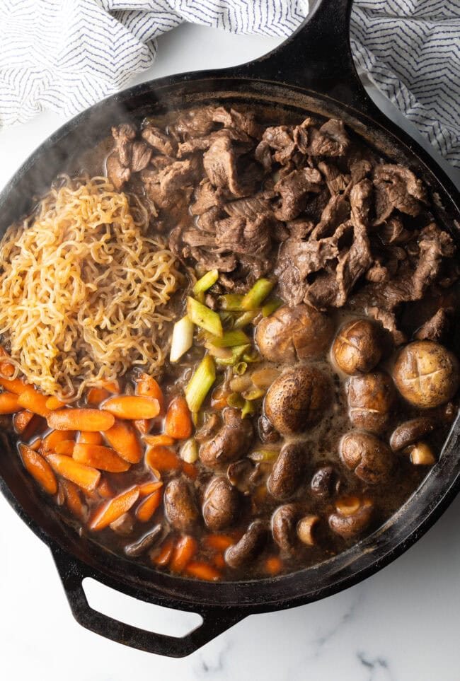 Black skillet with sections of noodles, mushrooms, beef, carrots, and beans.