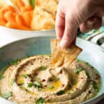 Hand using a pita chip to scoop eggplant dip from a light blue bowl.