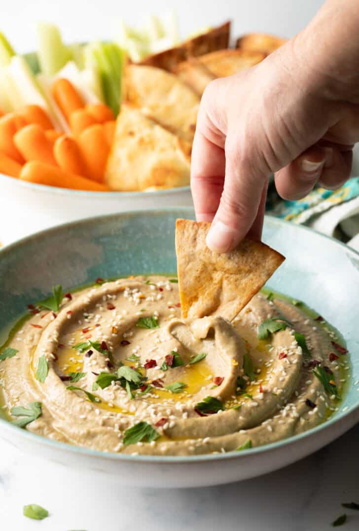 Hand using a pita chip to scoop eggplant dip from a light blue bowl.