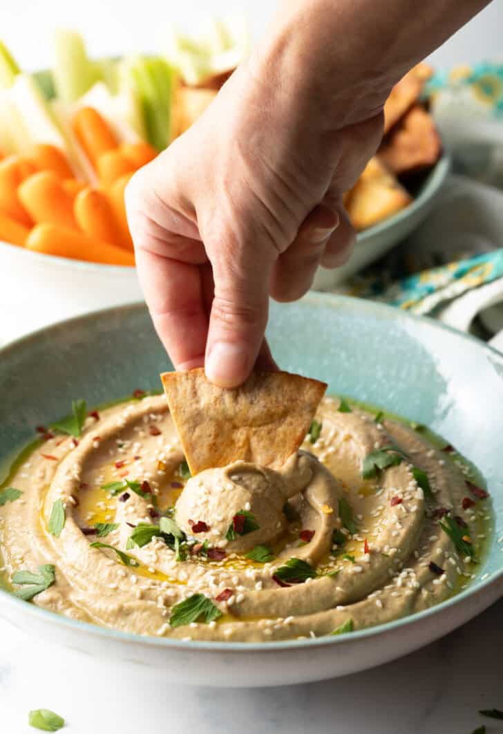 Hand using a pita chip to scoop eggplant dip from a light blue bowl.