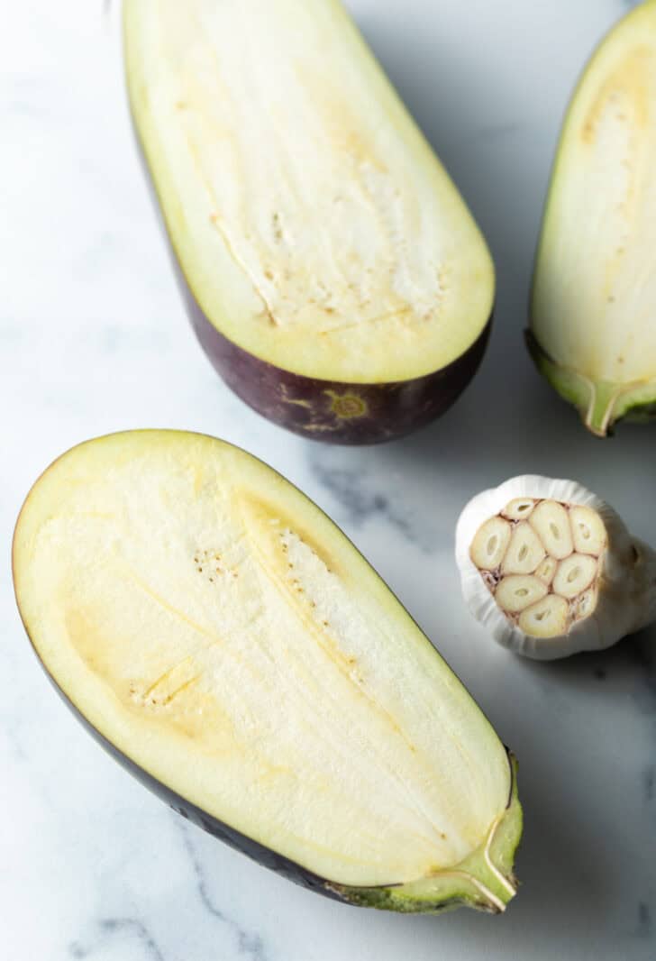 A raw eggplant halved, placed next to a head of garlic with the top chopped off.