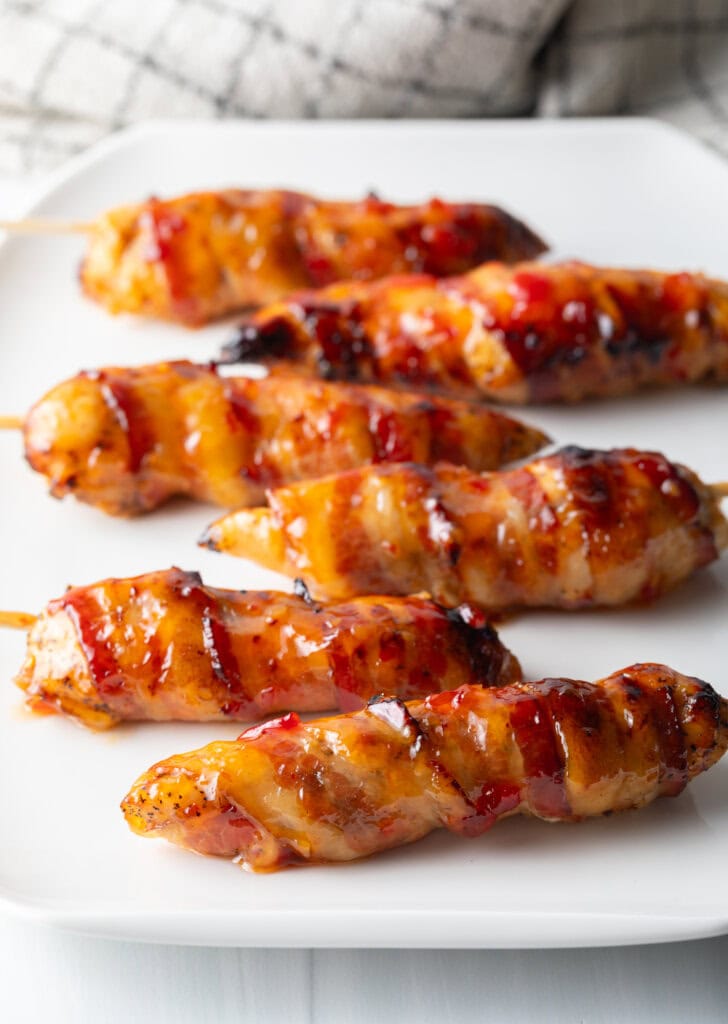 6 skewered bacon wrapped chicken tenders coated with hot sauce on a white plate.