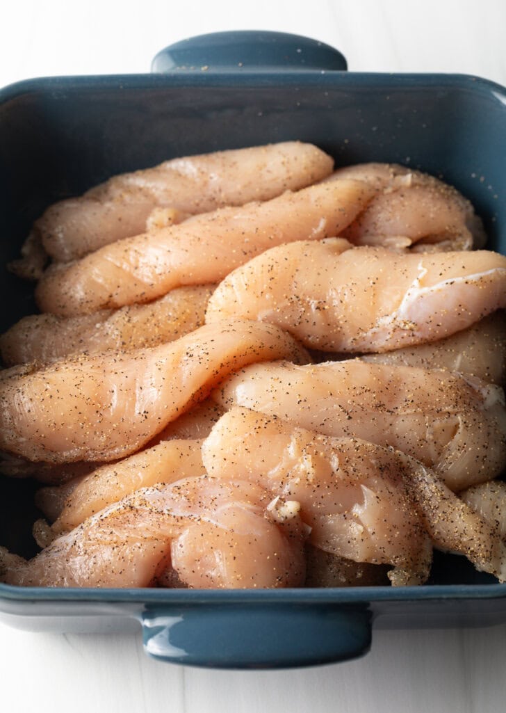 Square blue baking dish filled with raw chicken breast tenders.