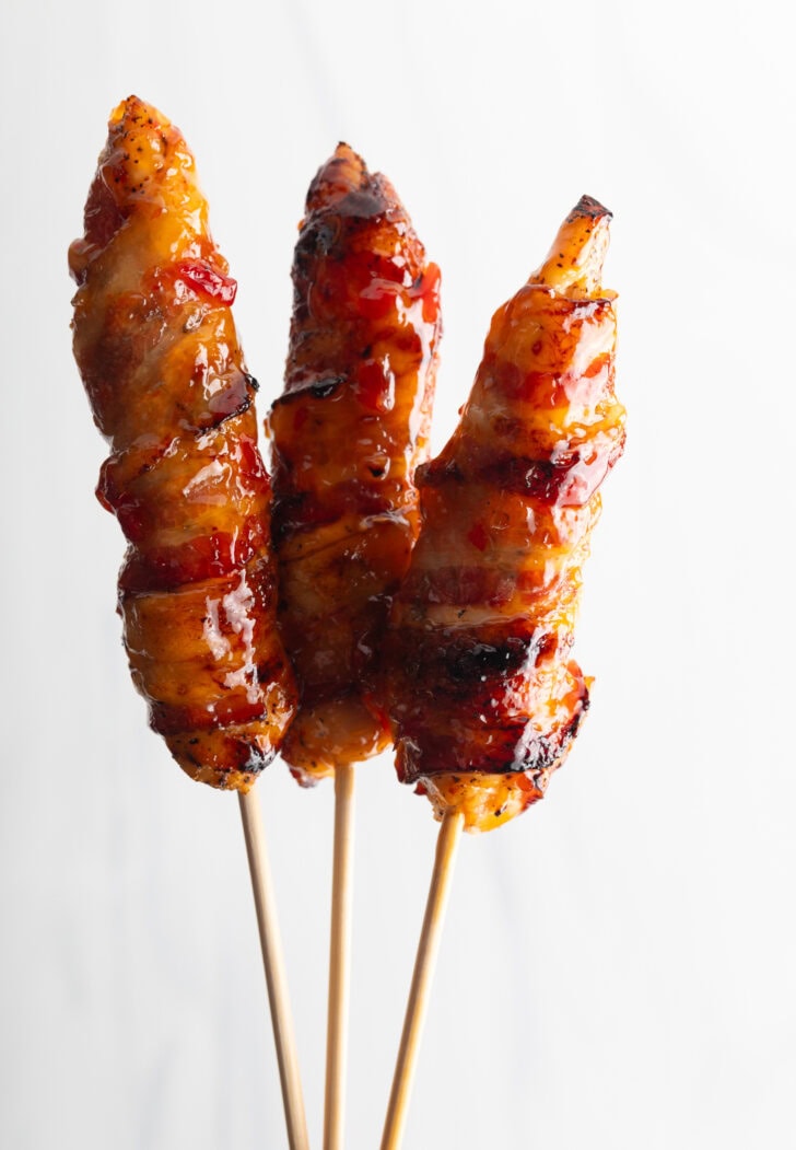 3 bacon wrapped hot sauce coated chicken skewers held up vertically to camera.