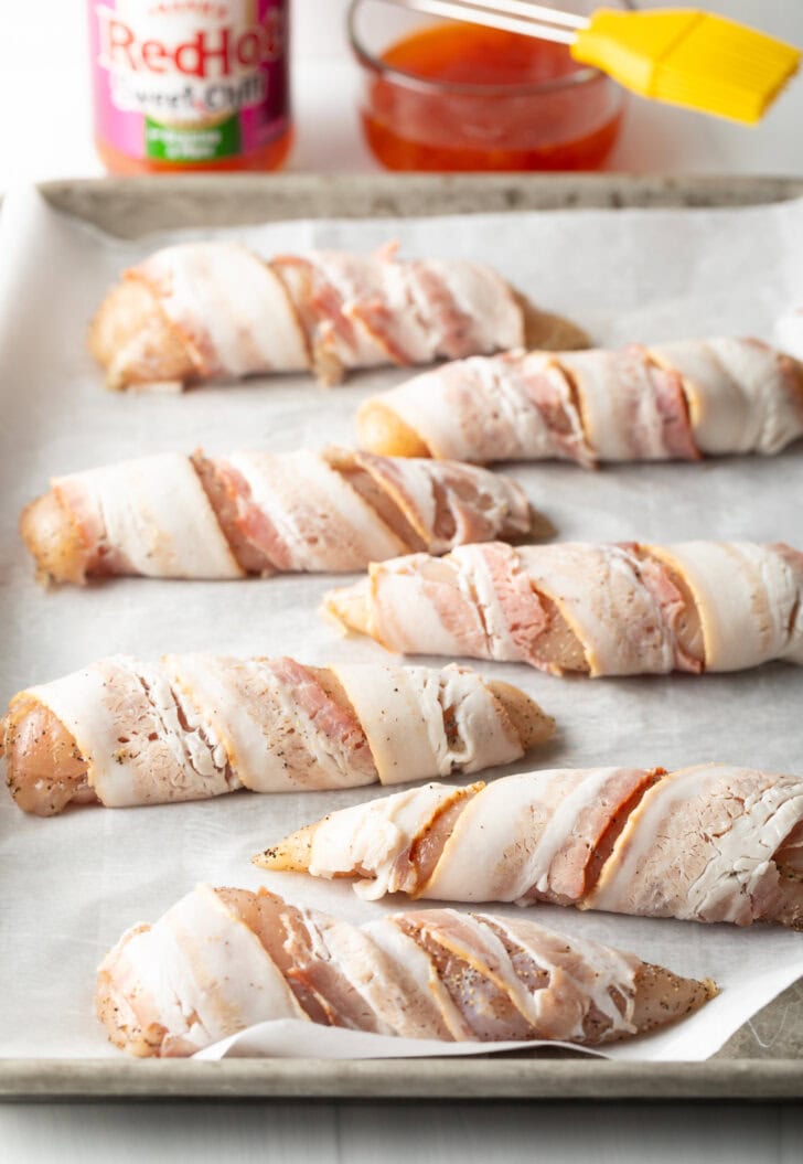 6 raw chicken breast tenders wrapped in raw bacon, placed on a baking sheet with white parchment paper.