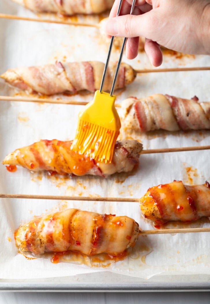 6 raw chicken tender wrapped in bacon on a baking sheet with parchment paper. A yellow basting brush is brushing on hot sauce.