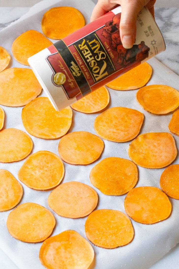 Hand sprinkling salt on top of sweet potato slices lined up on a baking sheet. 