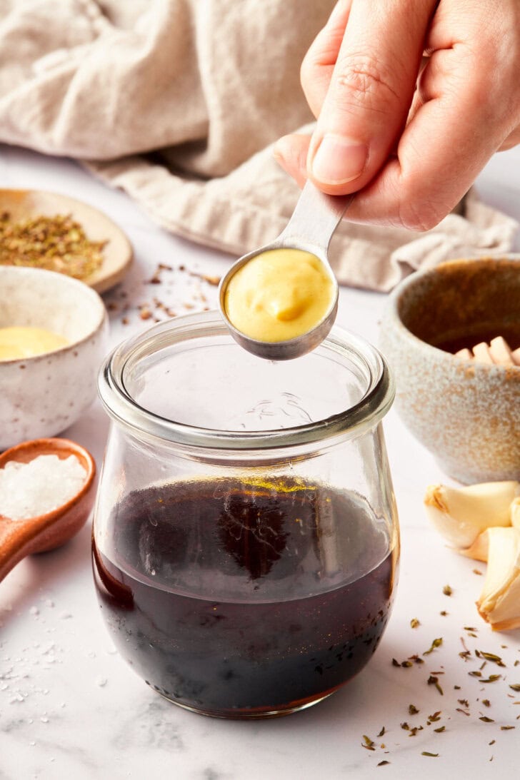 Balsamic Dressing Recipe - Hand holding a measuring spoon full of dijon mustard above a jar of balsamic vinegar. 
