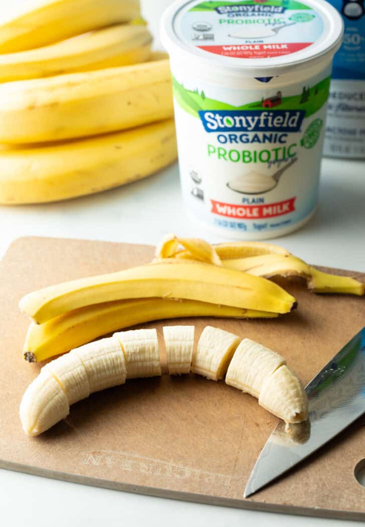 A banana is chopped on a cutting board, with peel and a carton of whole milk yogurt behind it.