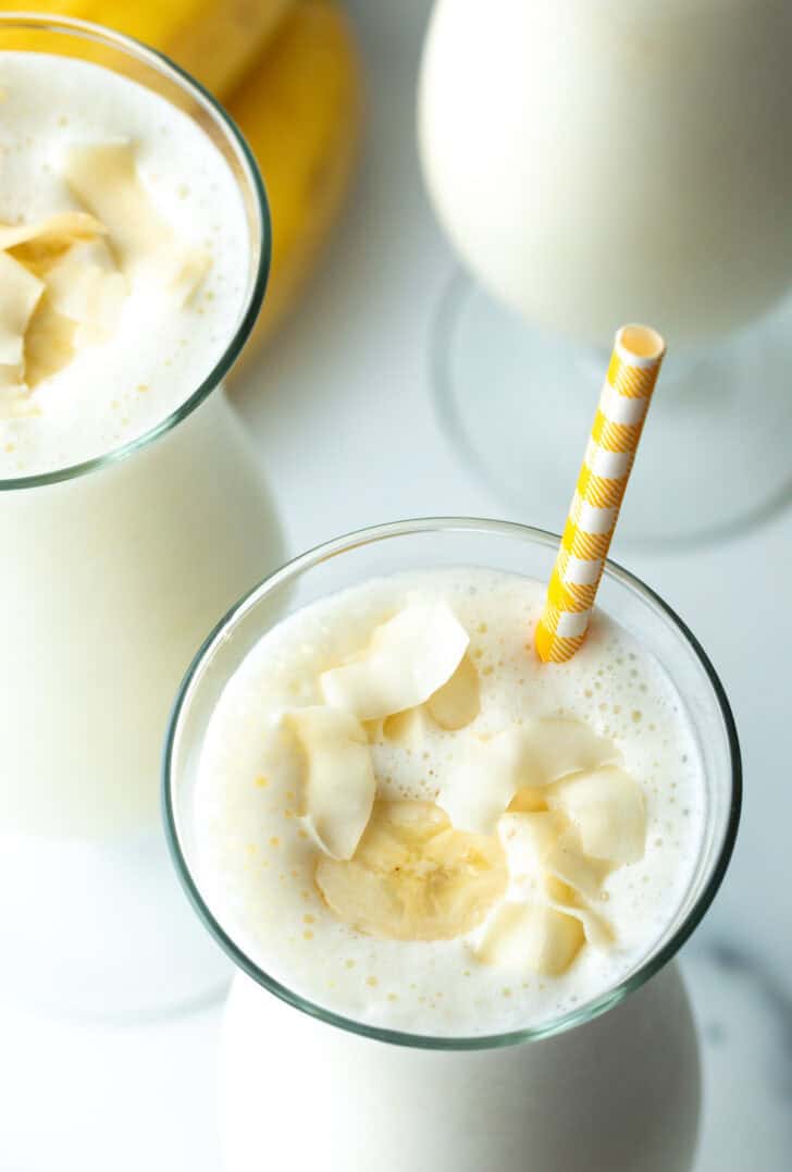 Top down view of a large mixed drink glass with banana smoothie and a yellow striped straw in each glass.